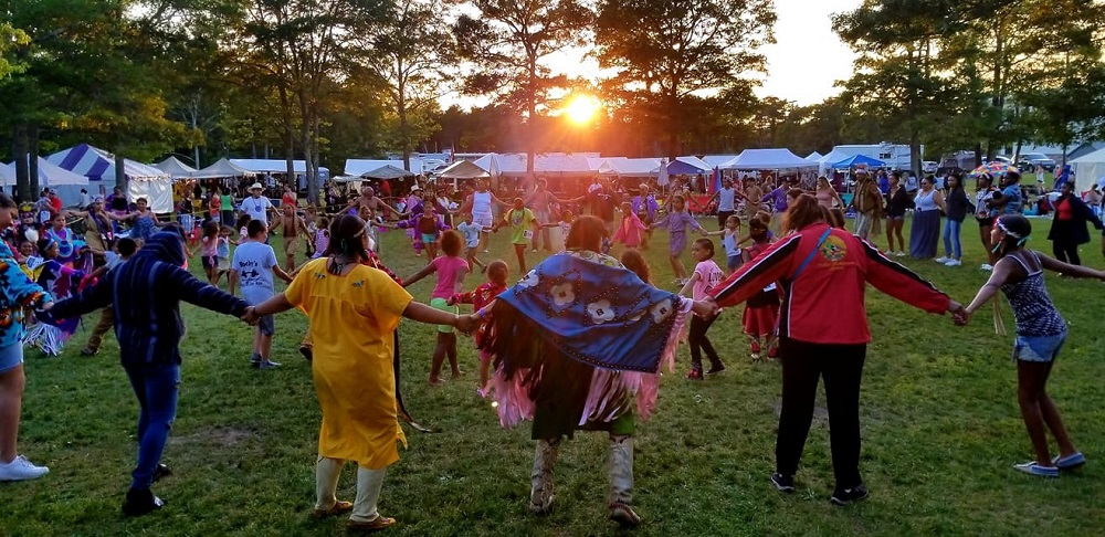Powwow of the Mashpee Wampanoag Tribe showing community members linking hands in concentric circles; viewers and tents in background.