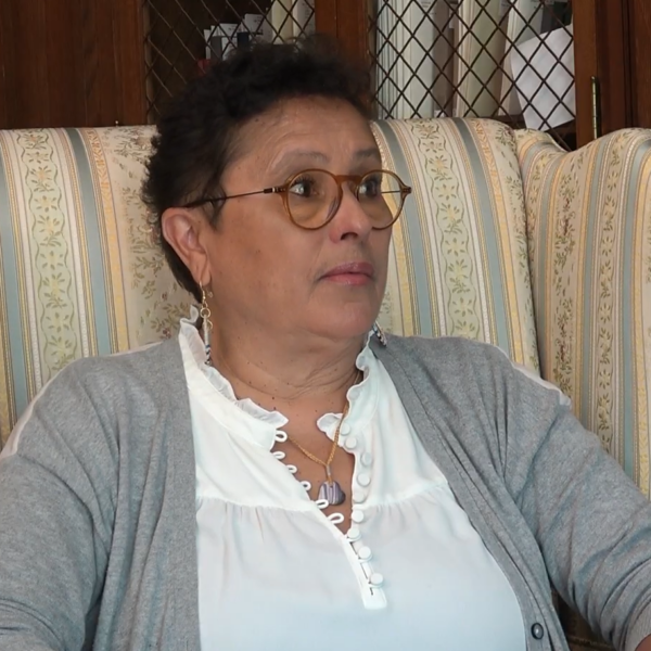 Paula Peters (Mashpee Wampanaog), wearing white blouse and grey cardigan, glasses, sits in armchair, bookcases and cabinets in background.