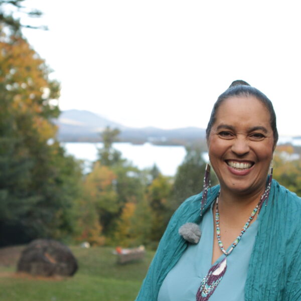 Narragansett woman smiling in blue blouse, blue-green shaw, beaded necklace and earrings; trees, hills, and shoreline in background.