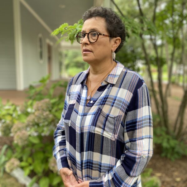 Paula Peters (Mashpee Wampanoag) in checkered blouse, stands at angle with hands interlocked looking towards the left; partial building and trees in background.